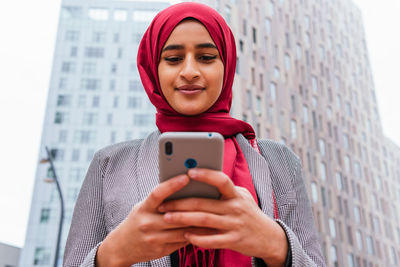 Low angle of positive ethnic female in hijab standing on street and chatting on social media with friends while using smartphone and enjoying weekend