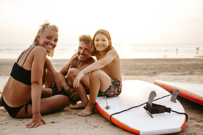 Portrait of happy family sitting with paddleboards at beach during sunset