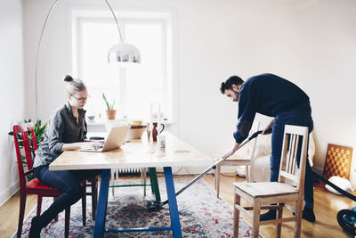 Woman using laptop while man cleaning carpet with vacuum cleaner at home