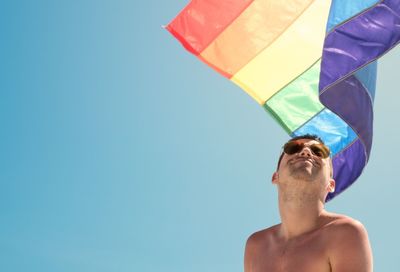 Low angle view of shirtless young man with rainbow flag against clear blue sky