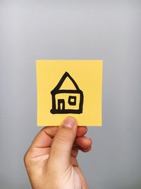 Cropped hand of person holding adhesive note with house symbol against gray background