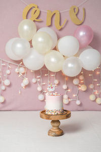 First birthday pink background - white and golden baloons with string lights, wooden cake stand