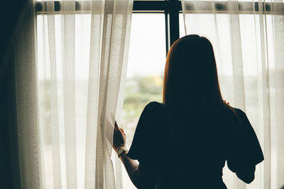 Rear view of woman looking through window at home