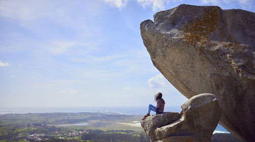 Side view of woman sitting on rock against sky