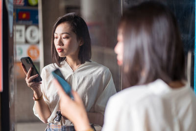 Serious ethnic woman browsing smartphone while standing on street and reflecting in mirror in taichung