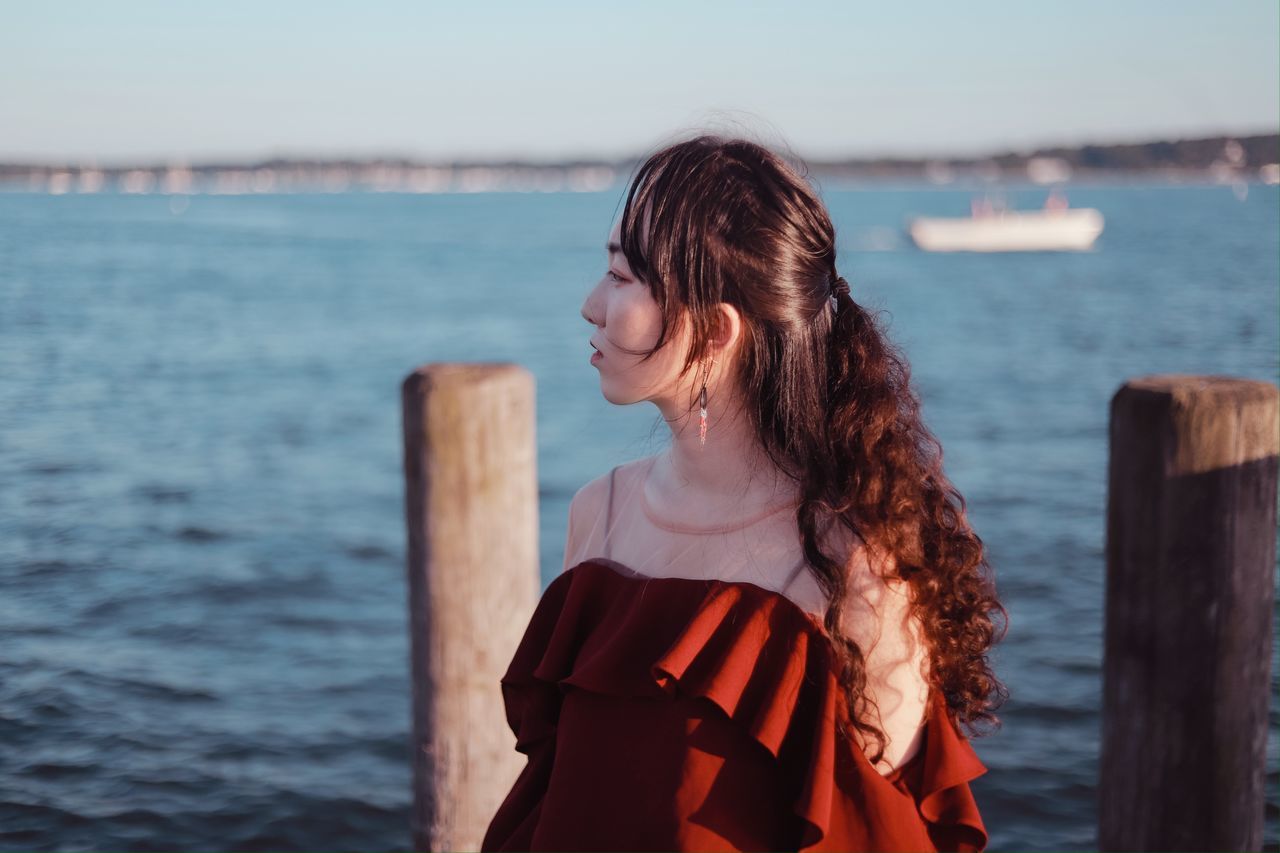 water, one person, hairstyle, sea, women, adult, young adult, nature, long hair, dress, sky, standing, clothing, brown hair, leisure activity, blue, fashion, focus on foreground, looking, waist up, wood, land, holiday, beauty in nature, vacation, lifestyles, trip, female, ocean, outdoors, casual clothing, tranquility, portrait, day, relaxation, looking away, redhead, beach, pier, smiling, emotion, person, contemplation, sunlight, travel, spring, photo shoot, tranquil scene