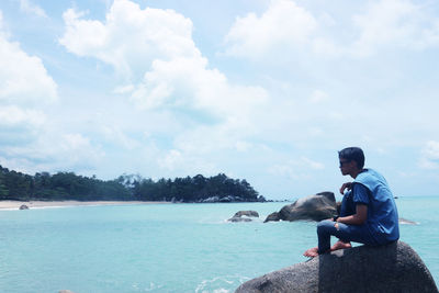 Side view of man sitting on cliff by sea against cloudy sky