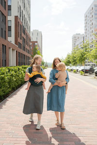 Full length of smiling caucasian mothers carrying their babies in slings on sunny day