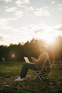 Full length of man with laptop using mobile phone while sitting on chair during sunset