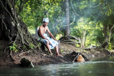 Full length of shirtless senior man sitting by tree at forest