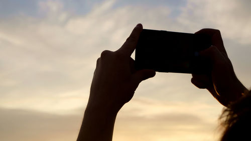 Low angle view of hand holding camera against sky during sunset