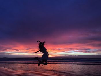 Full length of woman jumping at beach against cloudy sky during sunset