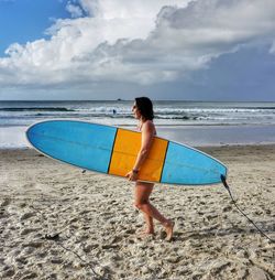 Side view of cheerful woman with surfboard walking at beach against cloudy sky