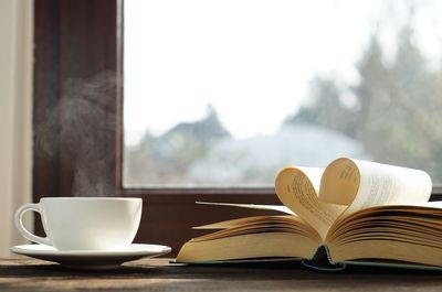 A cup of hot coffee with steaming next to heart shaped book on wooden table near by the window.
