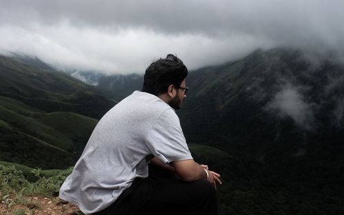Young man looking at view while sitting on mountain against sky