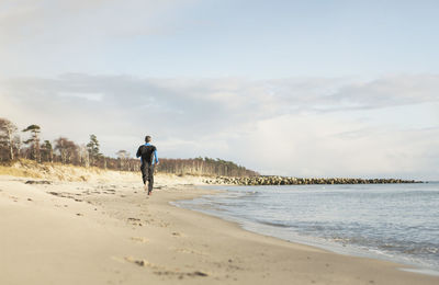 Rear view of man jogging on shore at beach against cloudy sky