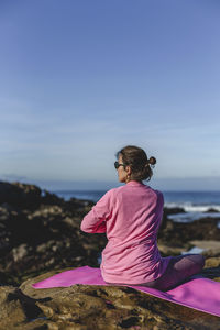 Rear view of mature woman exercising while sitting over mat against sky