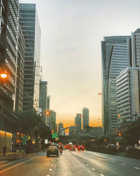 Traffic on city street by buildings against sky during sunset