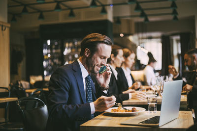 Smiling male business person talking on phone while eating food in restaurant