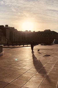 Silhouette people walking by buildings in city against sky during sunset