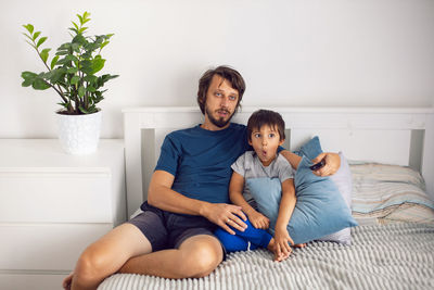 Bearded father and son are sitting on the bed in t-shirts and watching a football match on tv