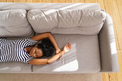 African american young woman wear stripped t-shirt resting, sleeping on couch with arms up at home