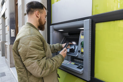 Young man with smart phone using atm