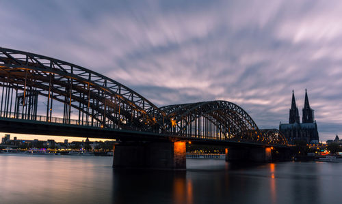 Illuminated bridge over river against cloudy sky