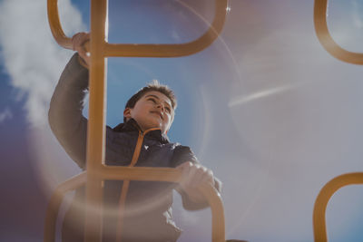 Low angle view of boy climbing jungle gym against sky at playground during sunny day