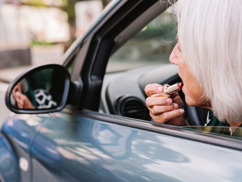 Side view elderly woman in elegant wear applying lipstick looking in rear view mirror while sitting on driver seat of modern car