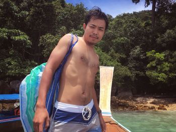 Portrait of shirtless young man standing in boat against trees