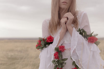 Midsection of young woman wearing white dress with artificial flowers while standing on field against sky