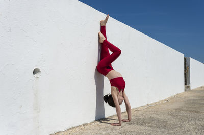Woman doing a handstand against a white wall with a blue sky wearing red.