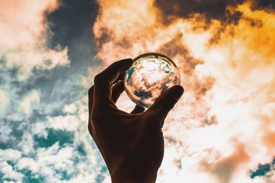 Cropped hand of man holding crystal ball against cloudy sky during sunset