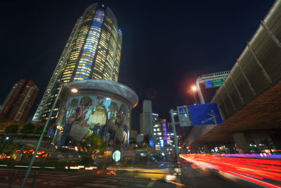 Light trails on road by buildings against sky at night