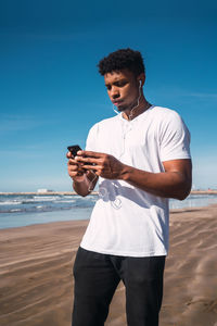 Young man using mobile phone while standing at beach against clear blue sky