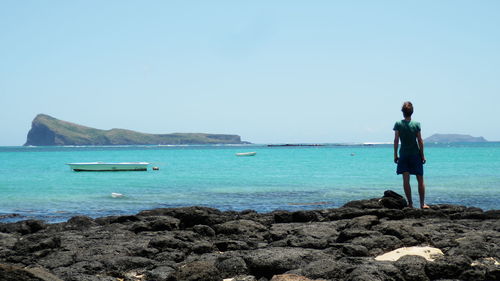 Rear view of man standing on rocks at beach against clear sky