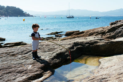 Side view of adorable child fisher in gumboots and overall holding small rod and fishing in shallow clear brook on rocks