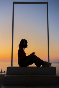 Silhouette woman sitting on shore against orange sky