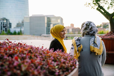 Delighted muslin female friends in hijabs sitting on bench and looking at each other while spending weekend in city