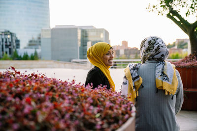 Delighted muslin female friends in hijabs sitting on bench and looking at each other while spending weekend in city