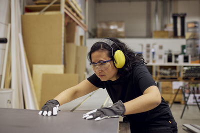 Serious female carpenter wearing protective eyewear and ear protectors while working in industry