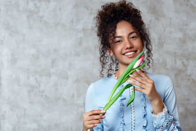 Portrait of smiling young woman holding tulip while standing against wall