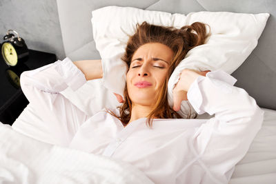 Young woman with covering ears with pillow on bed at home