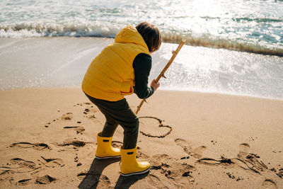 Boy in yellow rubber boots drawing heart shape on sand at the beach. kid child at autumn winter sea. 