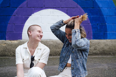 Happy woman tying hair with non-binary person sitting on footpath