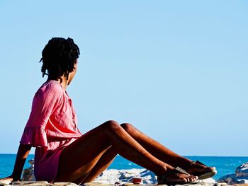 Woman sitting by sea against clear blue sky