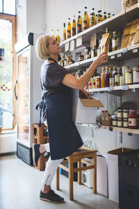 Full length of mature female employee arranging food products on shelf in deli