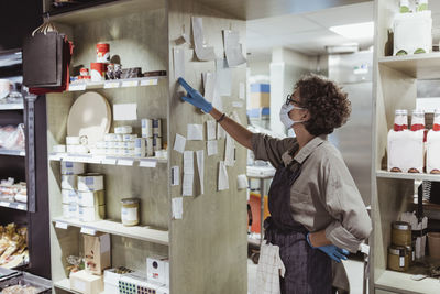 Female owner looking at adhesive notes stick on rack at delicatessen shop during pandemic