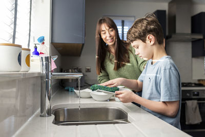 Happy woman with son washing dishes near kitchen sink